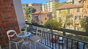 Un balcón con dos sillas blancas y una mesita, con vista a una calle con coches aparcados y edificios.