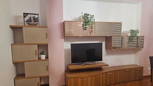 Una sala de estar con un televisor sobre un mueble de madera, una planta en un estante y otra planta en maceta en el suelo.