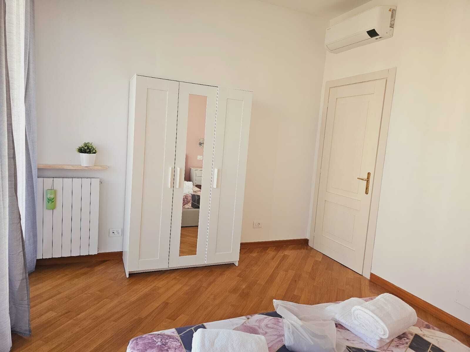 A bedroom with a wooden floor, a bed with a patterned bedspread, a white wardrobe, a small potted plant on a shelf, and a white door.