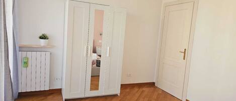 A bedroom with a wooden floor, a bed with a patterned bedspread, a white wardrobe, a small potted plant on a shelf, and a white door.