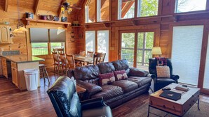 A cozy cabin interior with a wooden dining area, a leather sofa, and a kitchen with a large window.