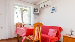 Et værelse med en rød sofa, en træstol, et spisebord dækket med en rød og hvidternet dug, et TV ophængt på væggen og en aircondition-enhed.
