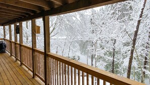 Eine Holzterrasse mit Geländer, die auf einen verschneiten Landschaftsraum und ein Gewässer blickt.