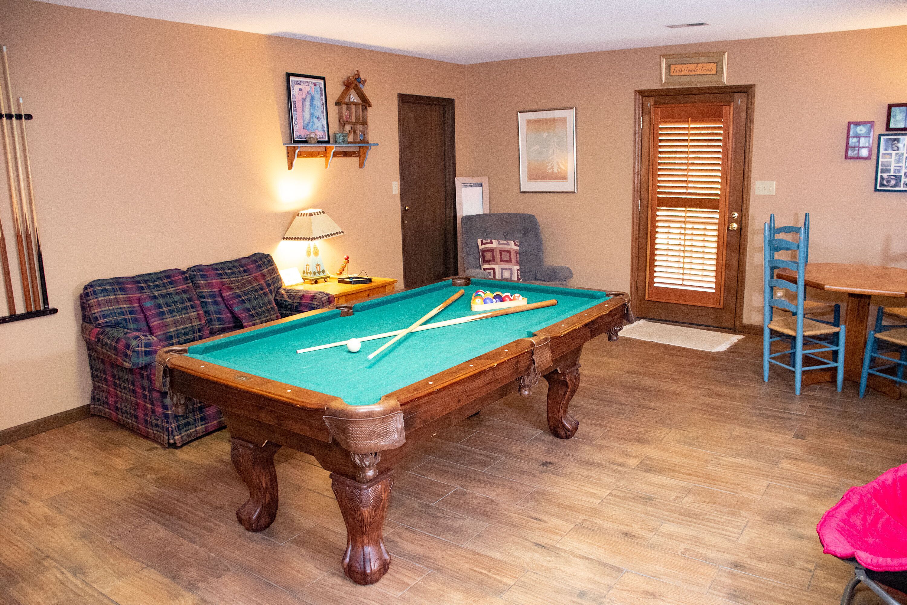 A pool table in a room with a wooden floor, a sofa, a lamp, and a door with a window.