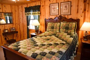 A wooden-paneled bedroom with a large bed, two bedside lamps, a nightstand, a mirror, and floral curtains. - Weeks Worth - Cabin in the Smokies (Sevierville)