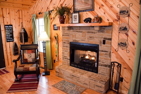 A cozy cabin interior with a stone fireplace, wooden walls, a rocking chair, and a fireplace mantel with decorative items.