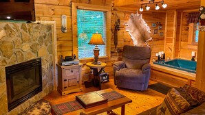 A cozy log cabin interior with a stone fireplace, a comfortable armchair, a wooden table, and a hot tub. - Sweet Serenity - Easy to Access Cabin By A Small Mountain Stream in Wears Valley (Sevierville)
