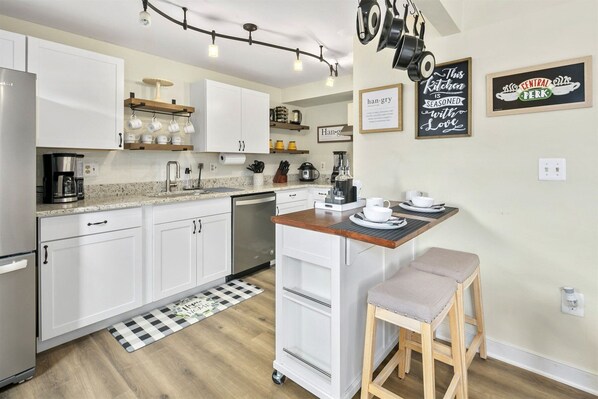Une cuisine moderne avec des meubles blancs, un îlot central en bois et un coin café. Les murs sont décorés et un tapis à carreaux se trouve à l’entrée.
