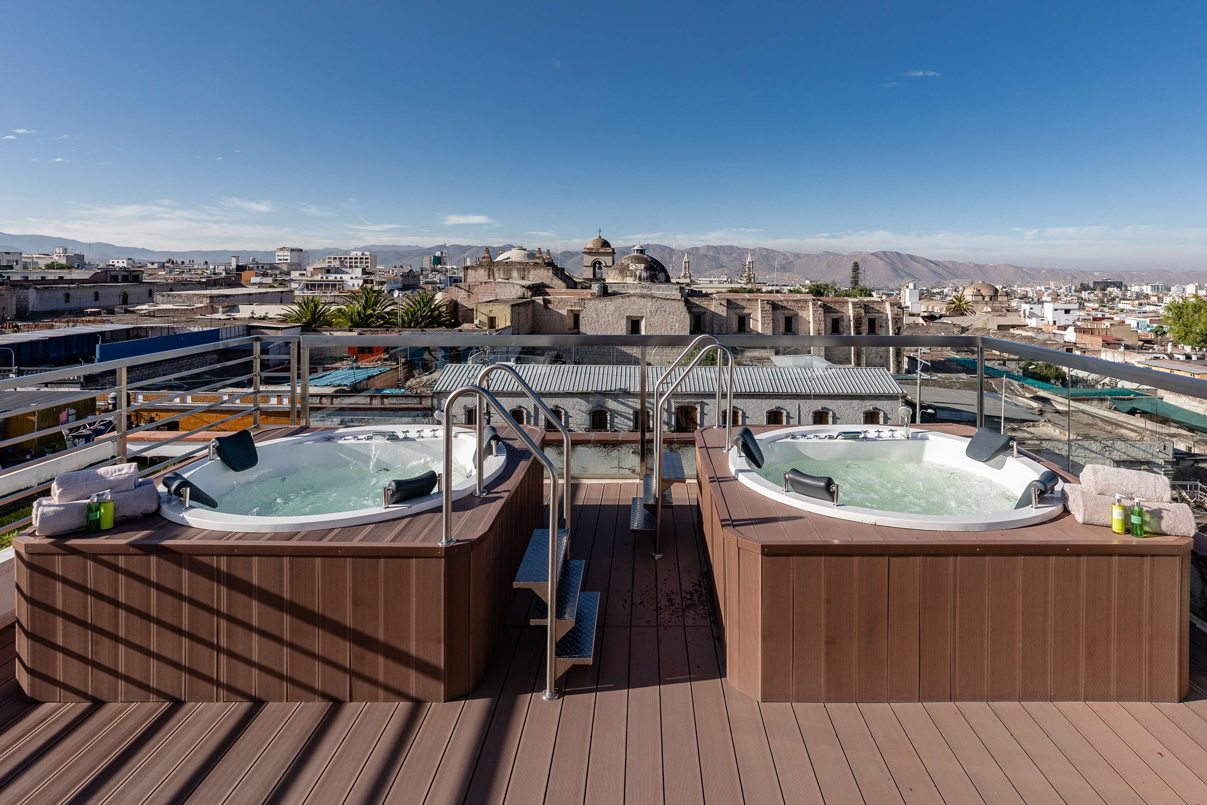 A rooftop deck with two hot tubs, wooden flooring, and a view of a cityscape with mountains in the background.