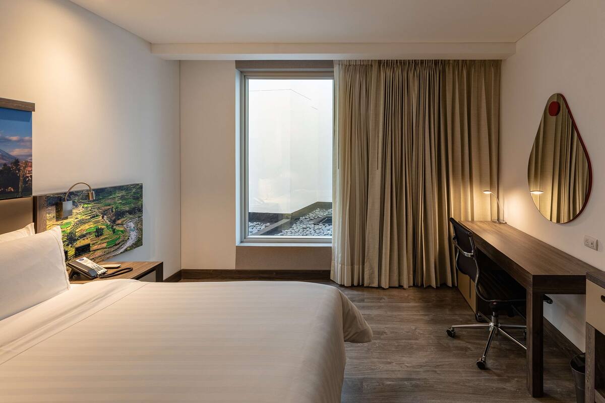 A hotel room with a bed, a desk, a chair, a window with curtains, a painting, and a mirror.