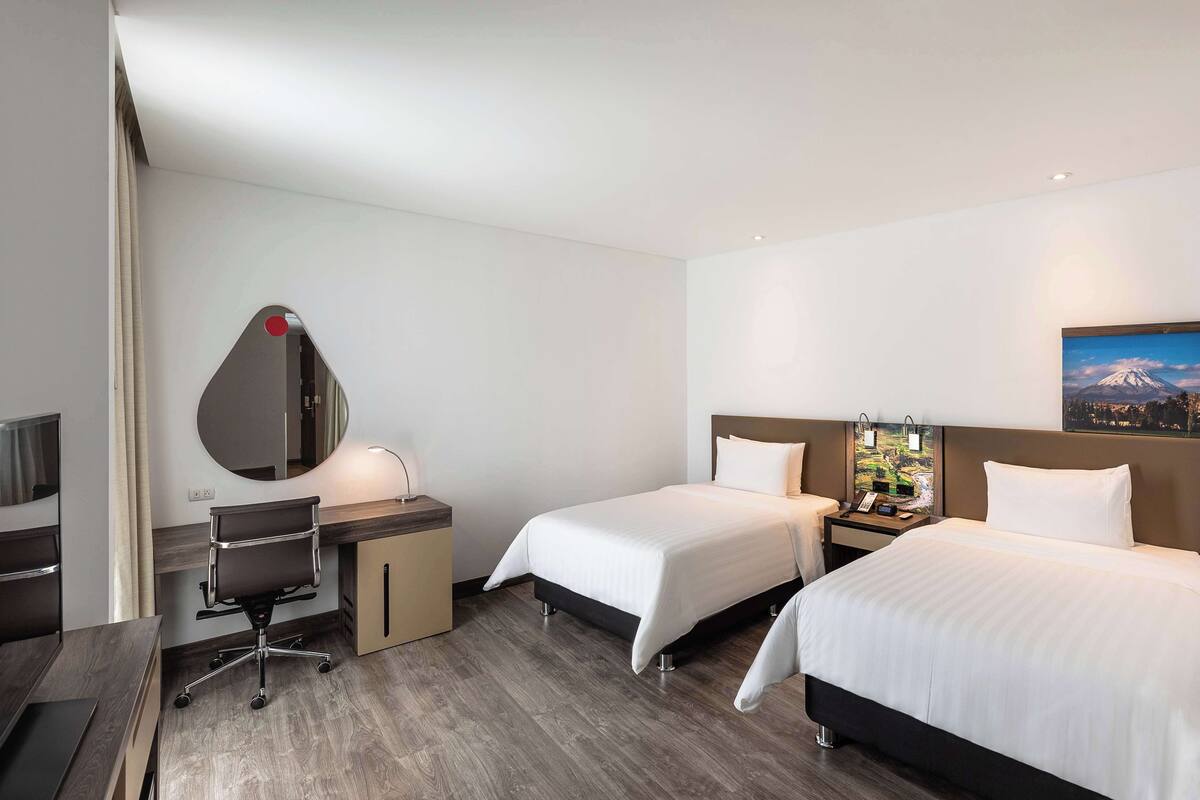 A hotel room with a desk, two beds, a mirror, a nightstand, and a painting on the wall.
