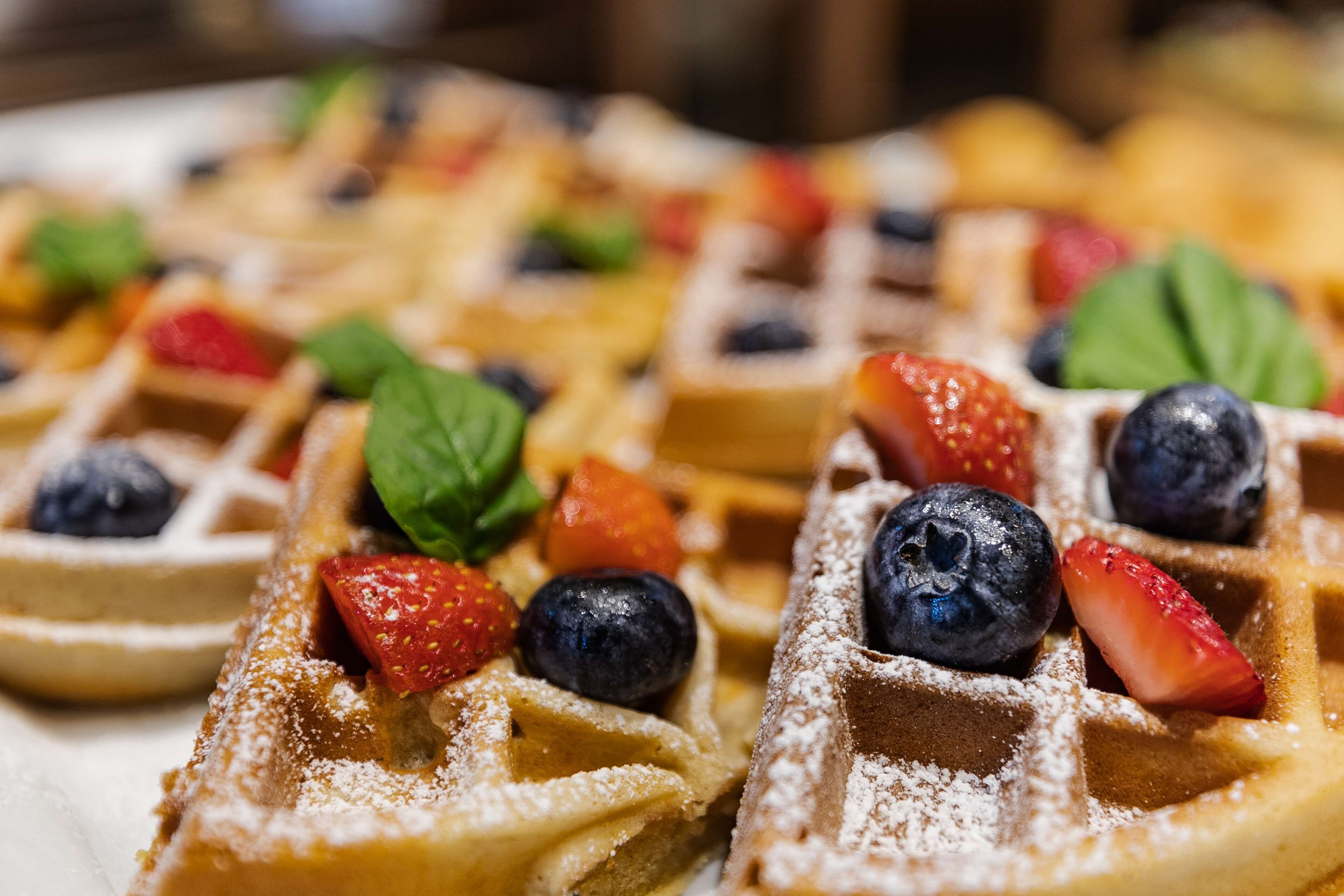 A close-up of a breakfast setting with waffles topped with strawberries, blueberries, and a dusting of powdered sugar.