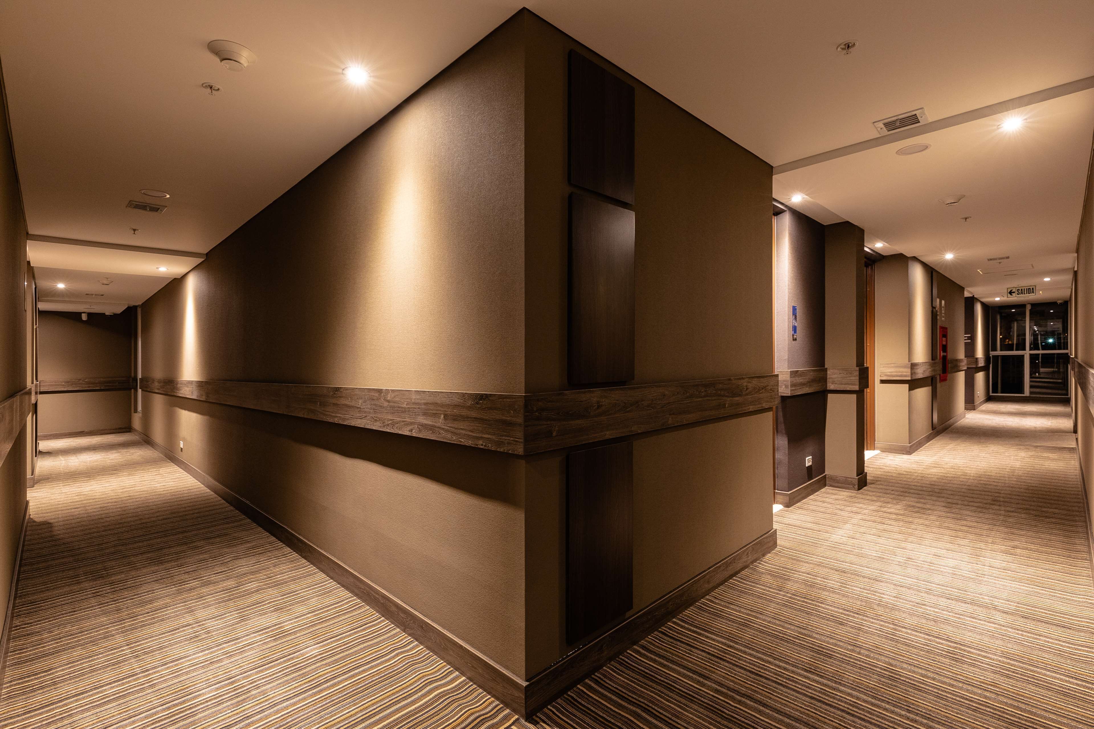 Un couloir d’hôtel au design moderne, avec un mur d’accent en bois, un éclairage encastré et un sol recouvert de moquette.
