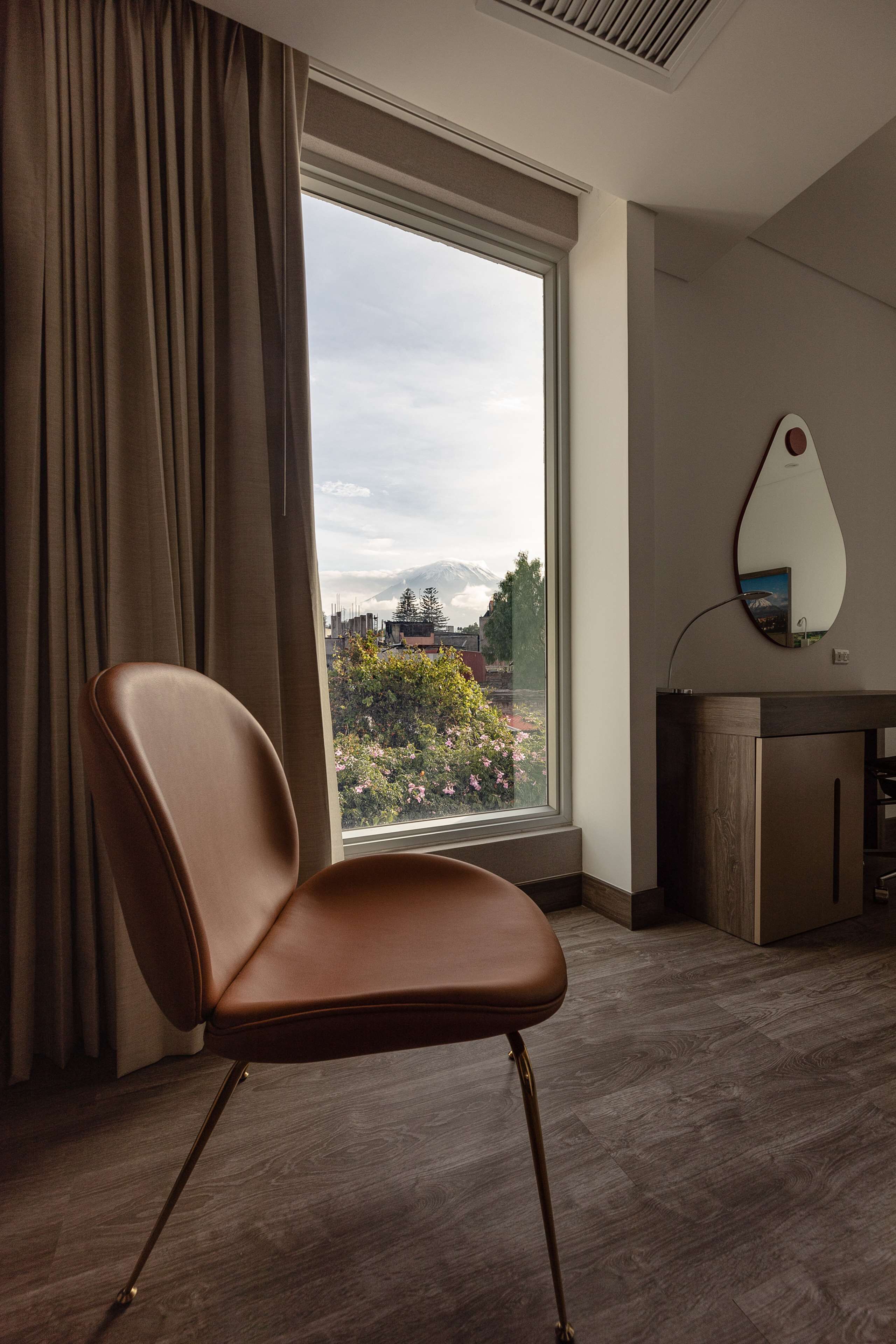 A modern room with a large window, a brown leather chair, a wooden desk, and a wall-mounted mirror.