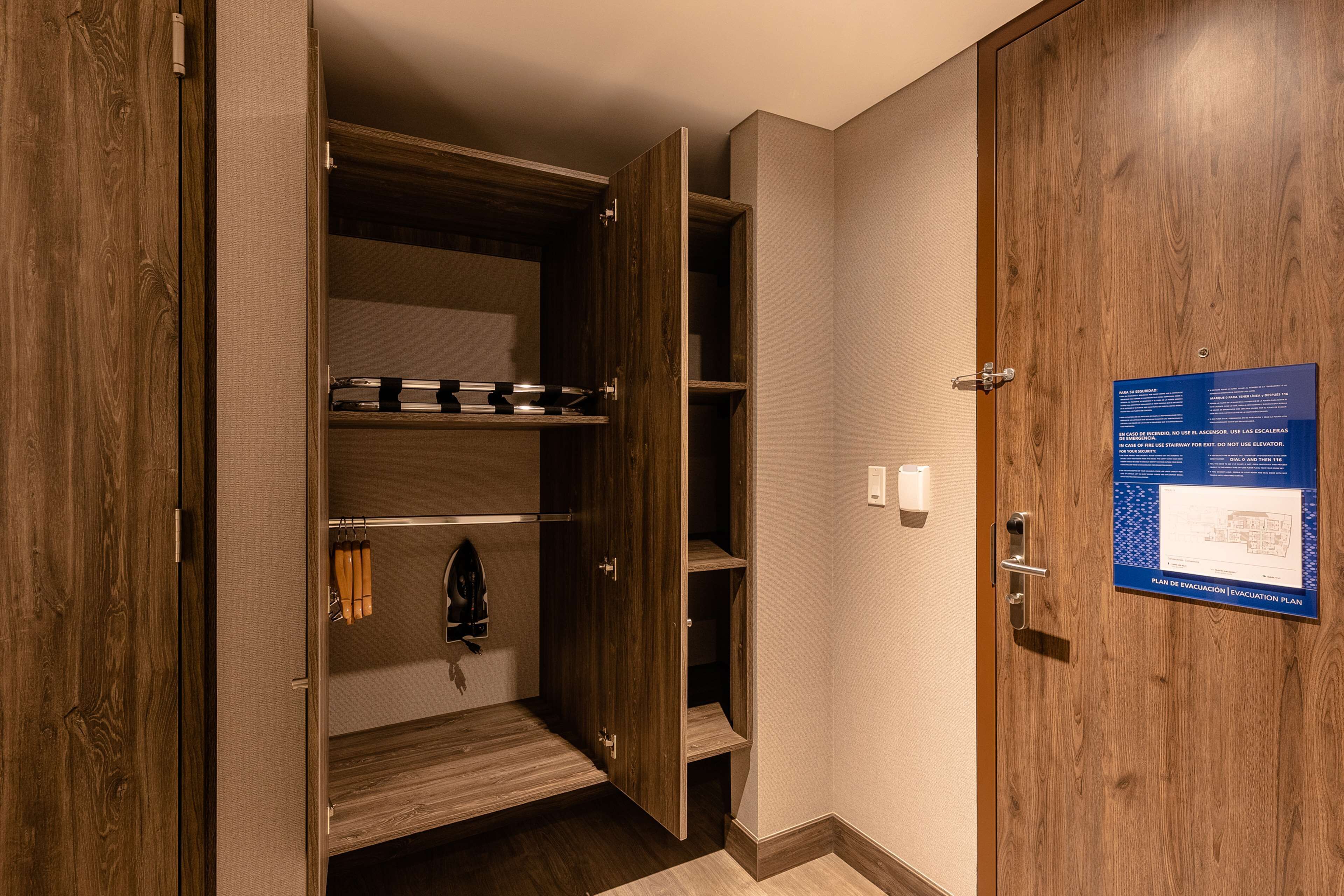 A hotel room with a wooden wardrobe, a blue evacuation plan poster, and a coat hanging on the door.