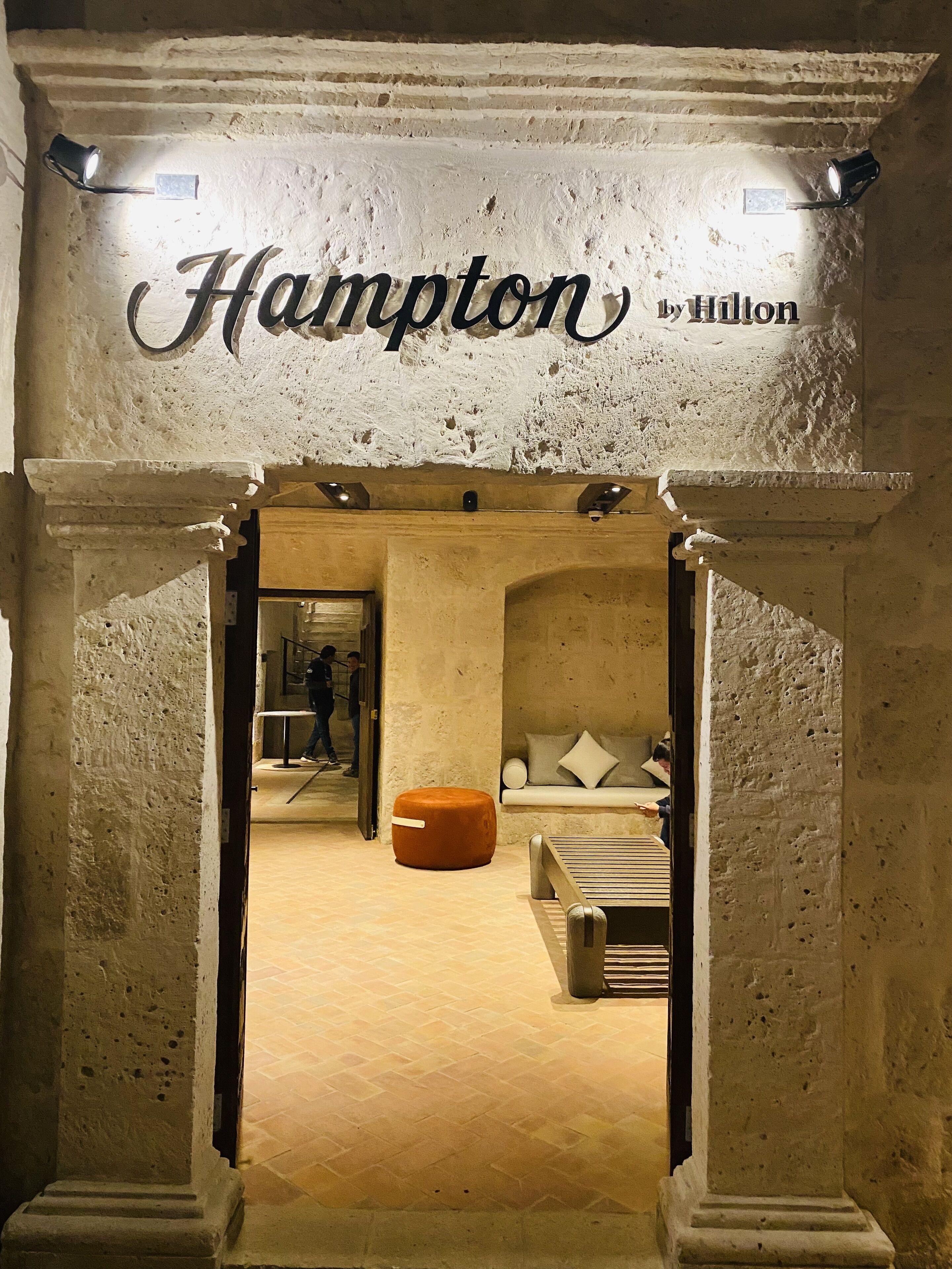 Ein Eingang zum Hampton by Hilton mit einer Steinfassade, einem Schild über der Tür und einem Blick in den Innenbereich, der eine Couch und eine hölzerne Bank zeigt.