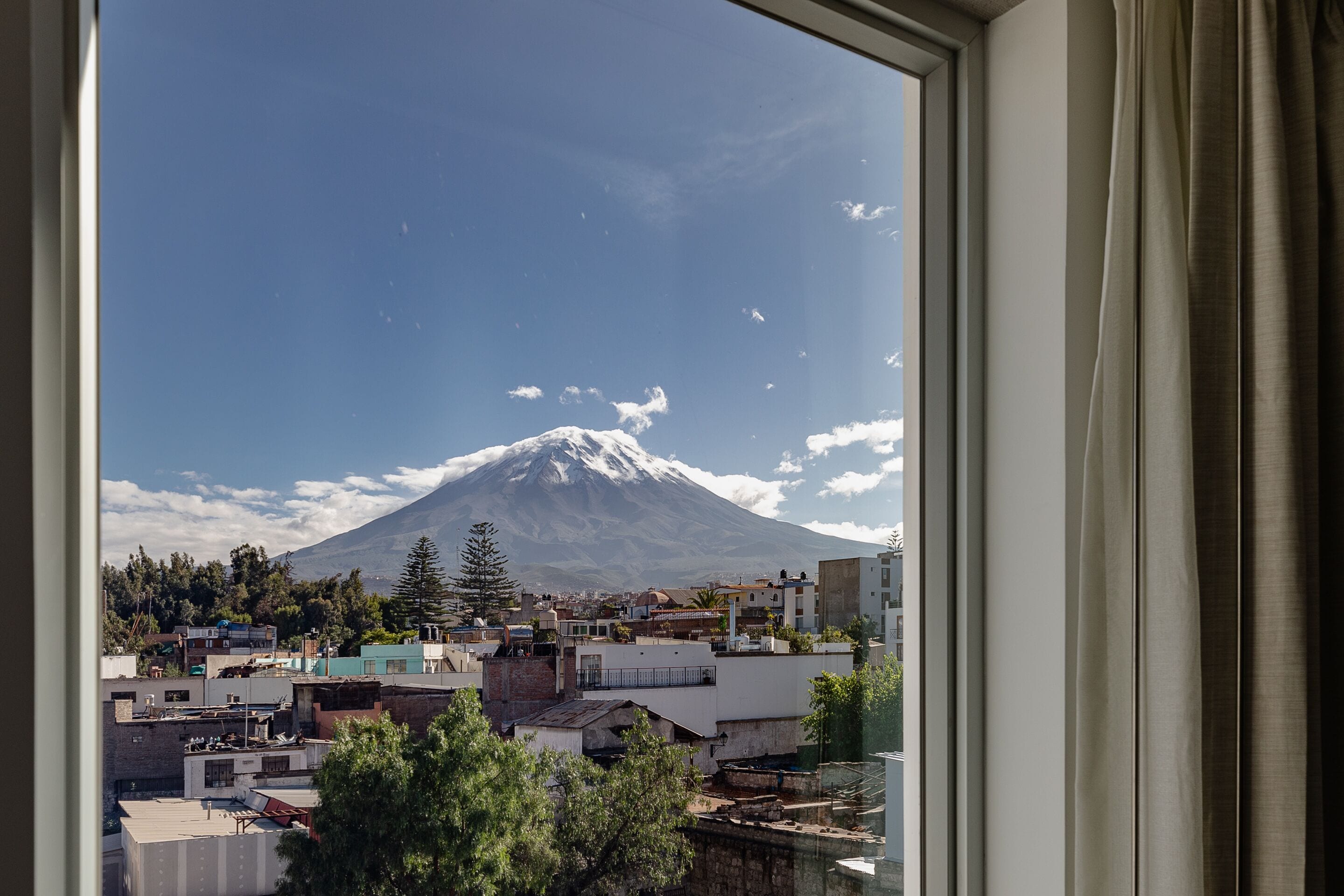 A view of a snow-capped mountain through a window, with buildings and trees in the foreground.
