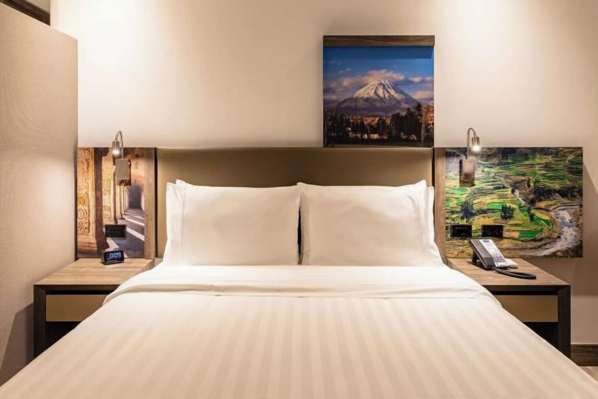 Habitación de hotel con una cama grande, dos mesitas de noche, una mesita con lámpara y obras de arte en las paredes.
