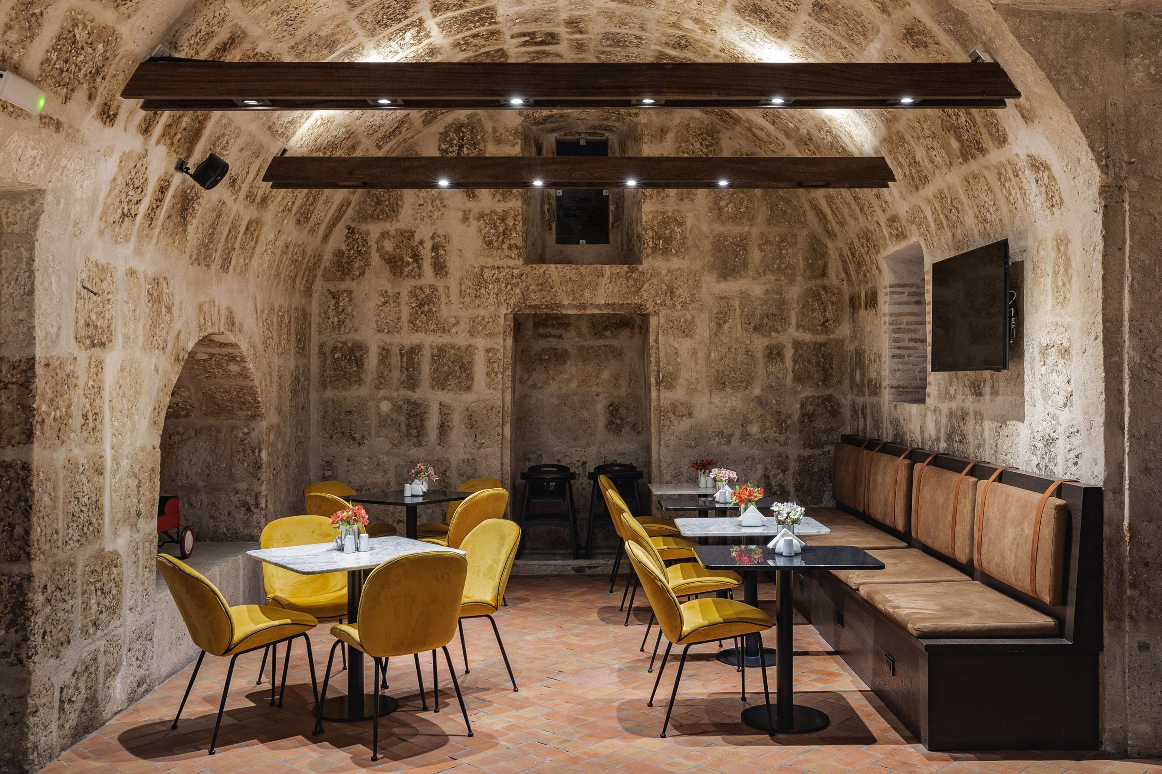 A restaurant with stone walls, yellow chairs, and a long bench. Tables are set with flowers.