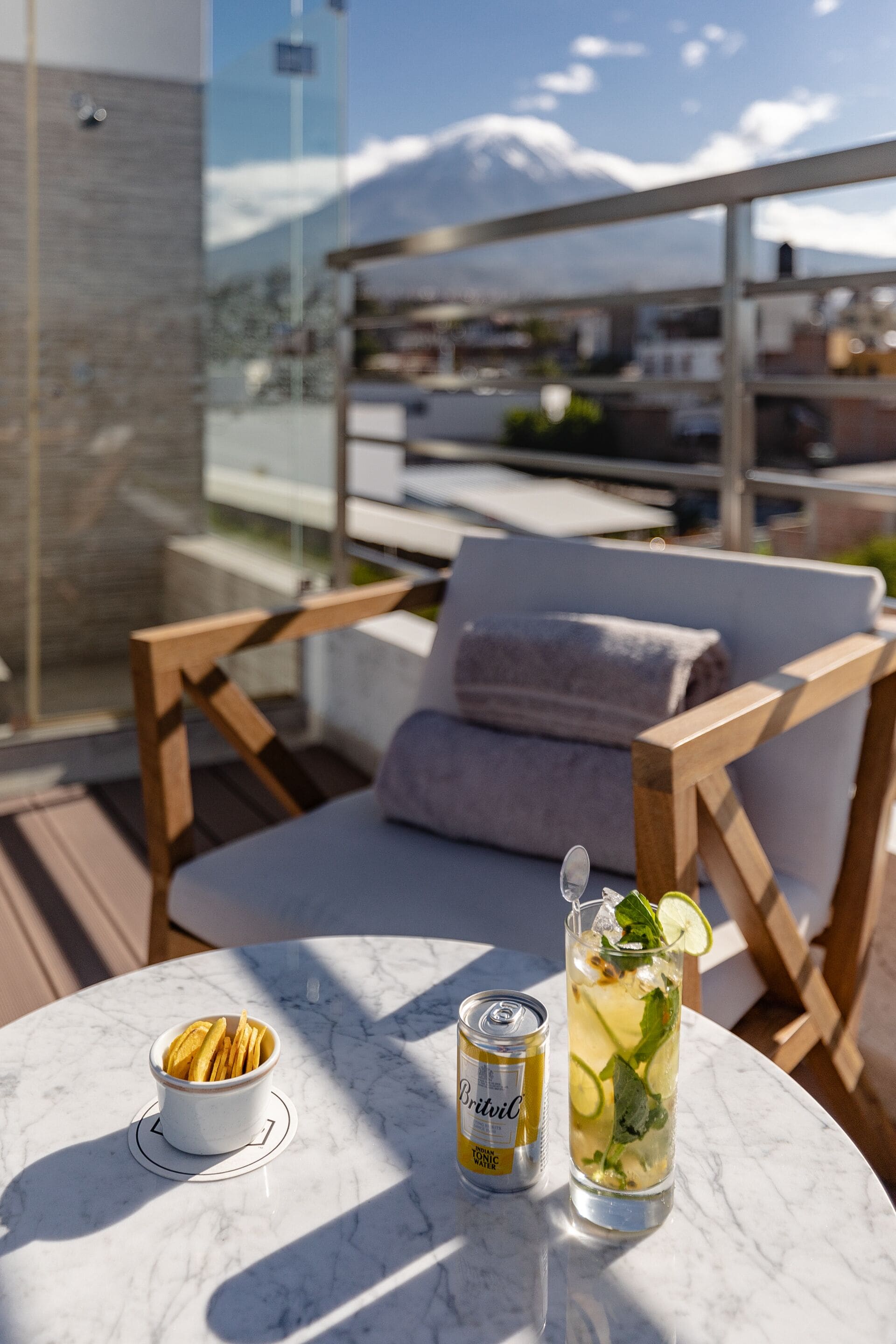 Ein Tisch mit einer Dose Britvic Tonic Water, einem Glas eisgekühlten Getränk mit Limette und Minze sowie einer Schüssel Chips im Freien.