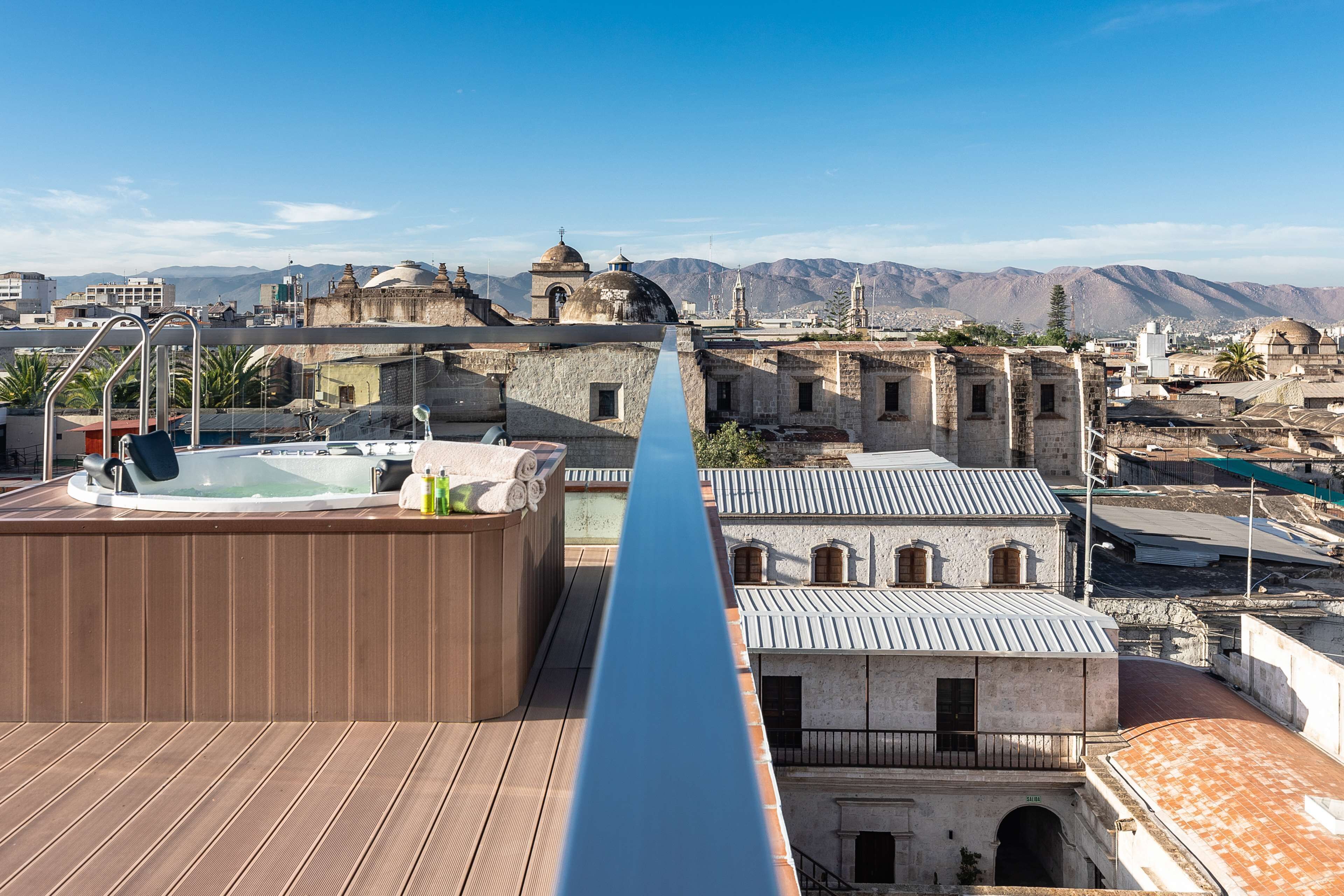 Una terraza en la azotea con un jacuzzi y cubierta de madera, con vistas a un pueblo histórico de arquitectura tradicional y montañas al fondo.