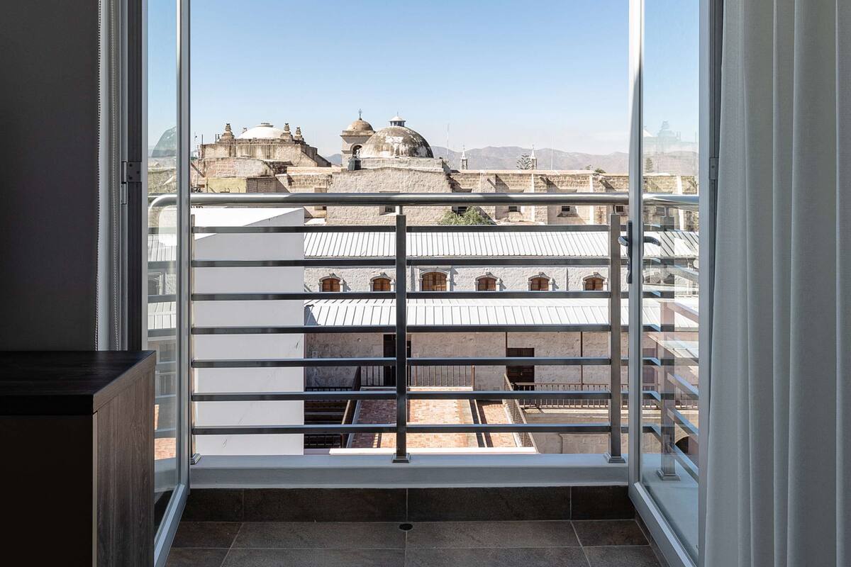 A room with a large window, a balcony with a metal railing, and a view of buildings in the distance.