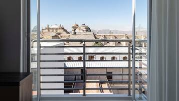 Una habitación con un ventanal grande, un balcón con barandilla metálica y vistas a edificios en la distancia.