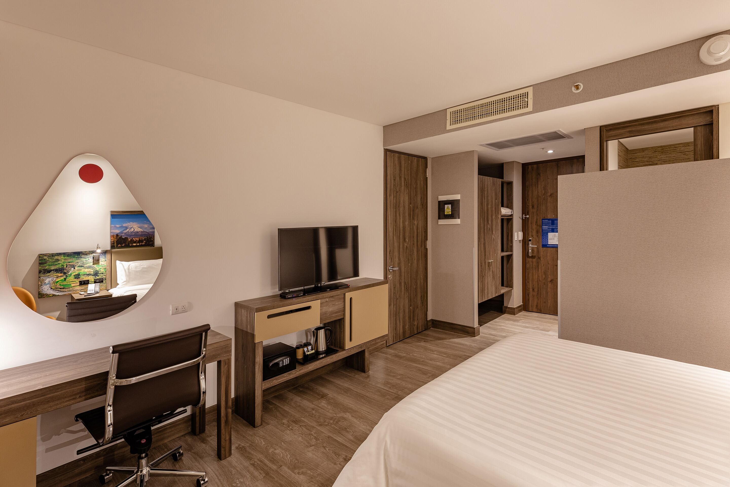 Una habitación de hotel con cama, escritorio con silla, televisor, espejo y armario.