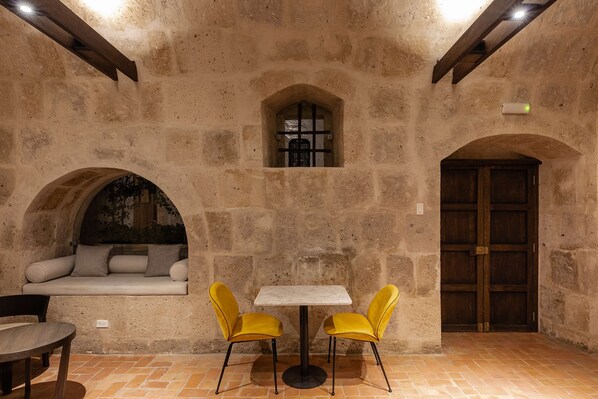 Una habitación con paredes de piedra, una mesa redonda, dos sillas amarillas, un banco con cojines y una puerta de madera.