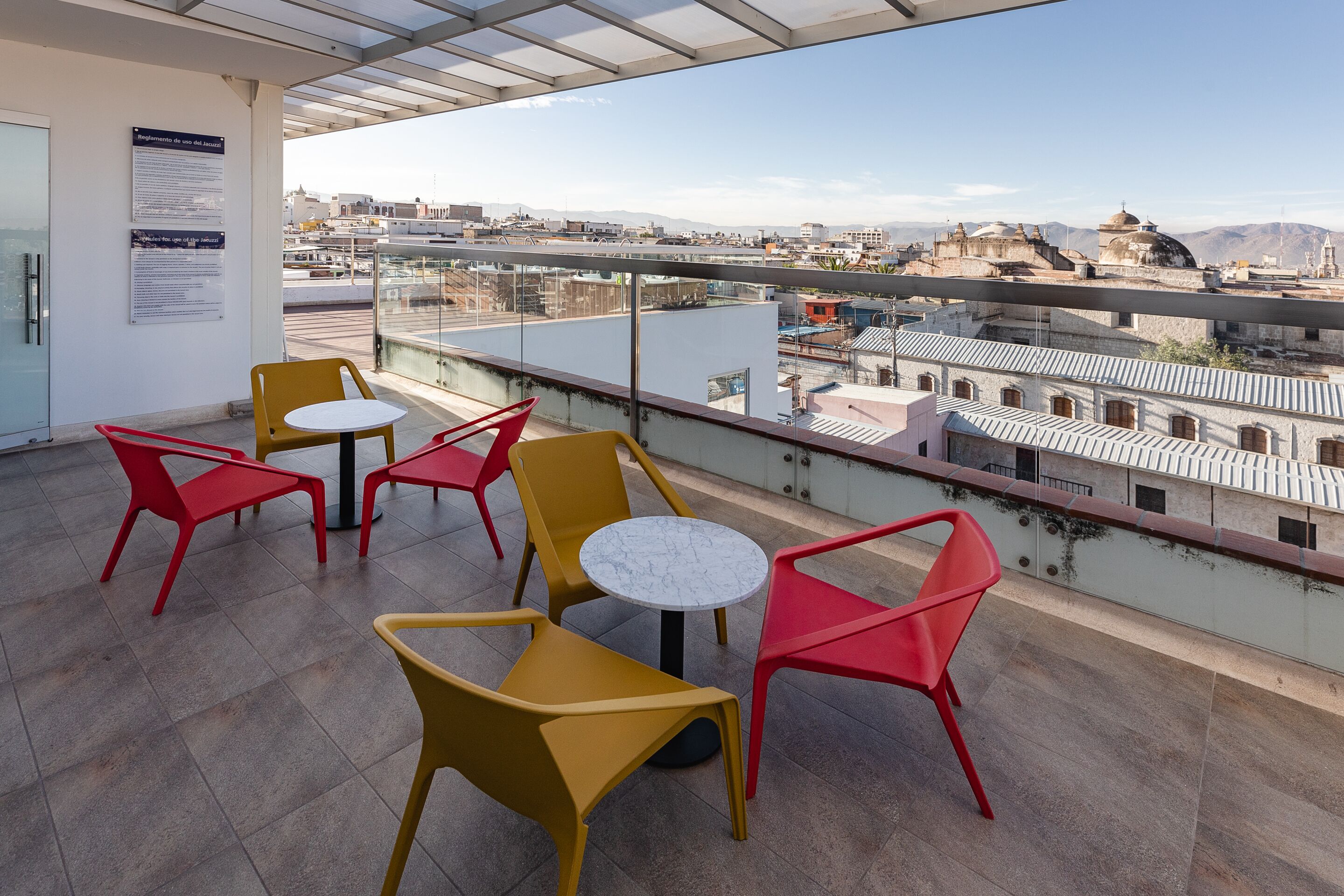 Un balcón con sillas amarillas y rojas, una mesa redonda pequeña y vista de un paisaje urbano con edificios y montañas al fondo.