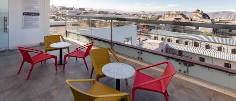 Un balcón con sillas amarillas y rojas, una mesa redonda pequeña y vista de un paisaje urbano con edificios y montañas al fondo.