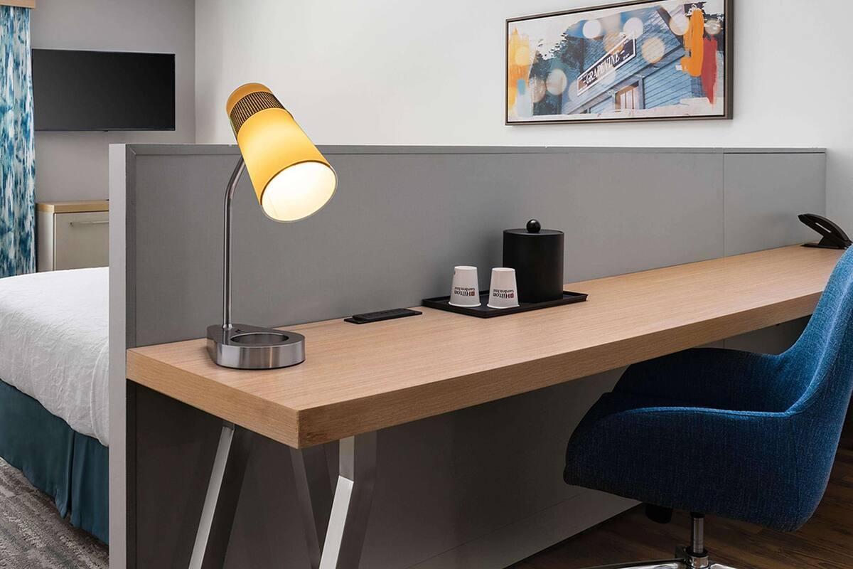 A hotel room with a wooden desk, a blue chair, a desk lamp, a black telephone, and a framed picture on the wall.