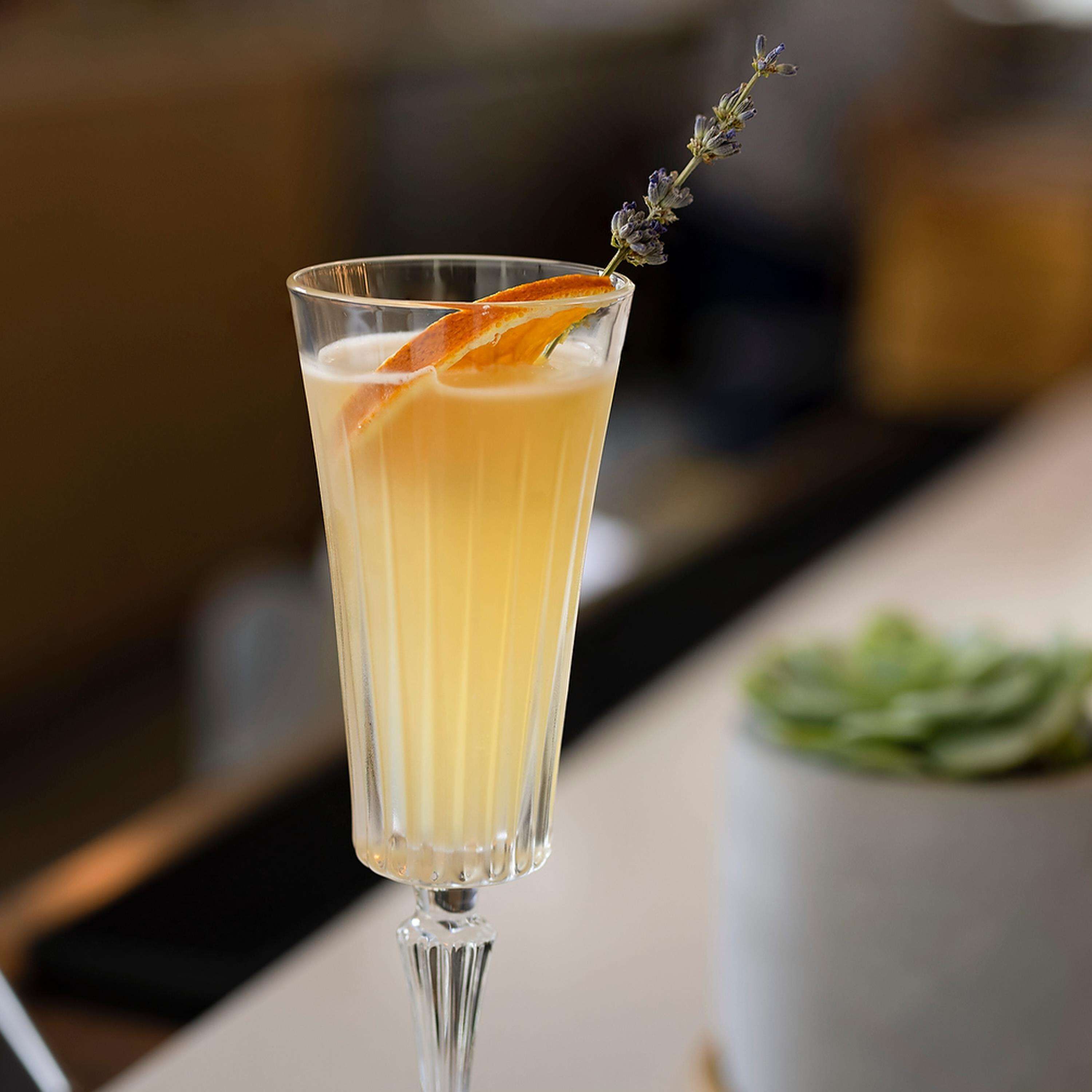A tall glass with a layered drink, garnished with an orange peel and a sprig of lavender.