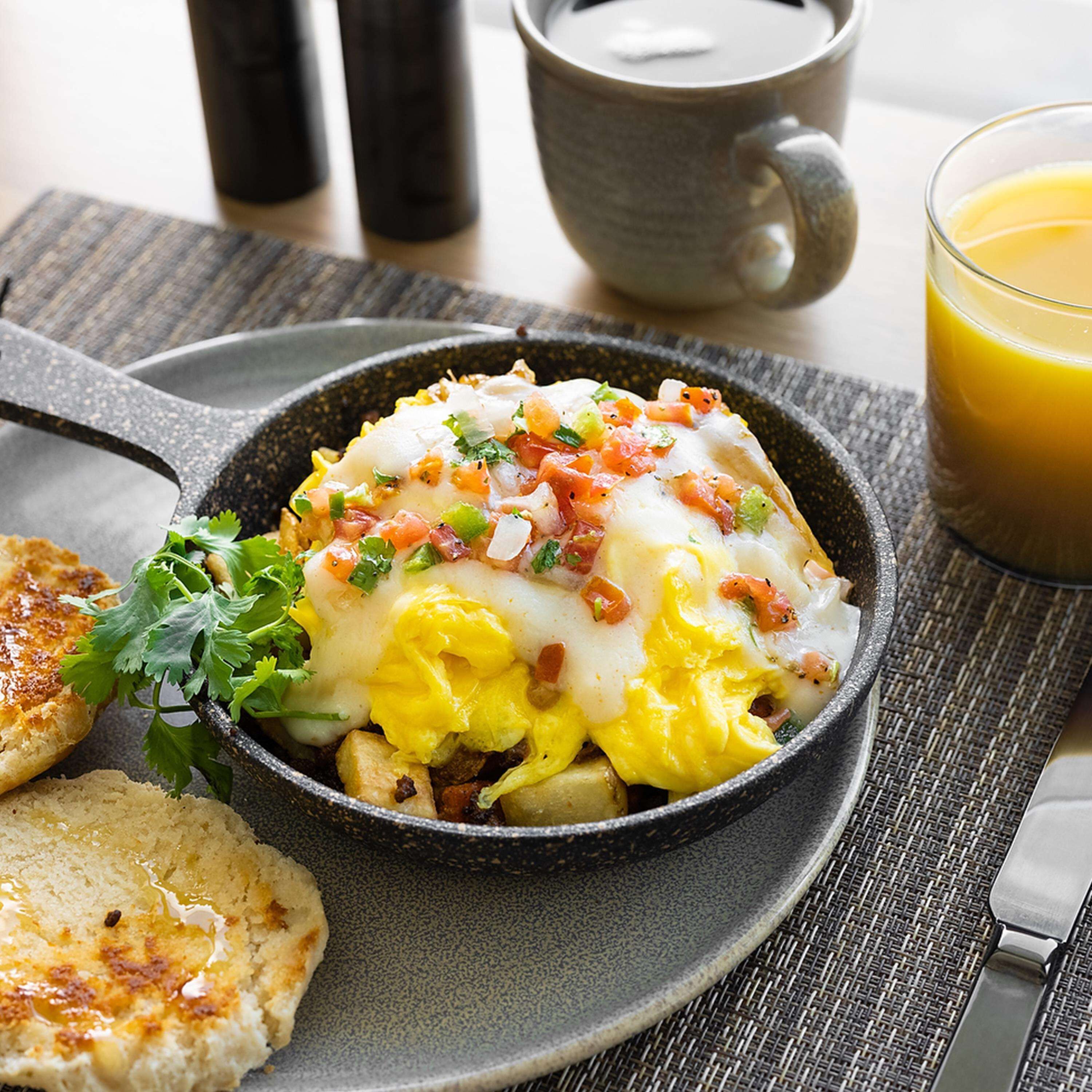 A breakfast dish with eggs, cheese, and vegetables in a black bowl, accompanied by toast and a glass of orange juice.
