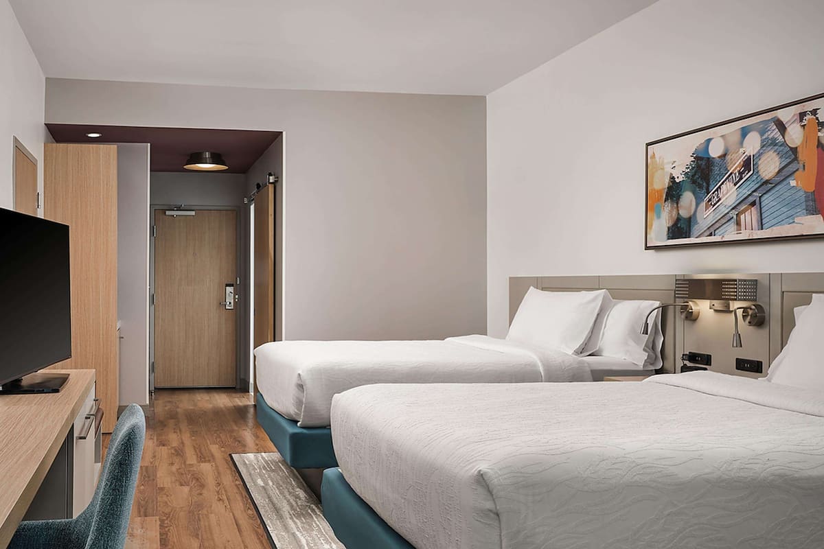 A hotel room with two beds, a desk, a chair, and a painting on the wall.