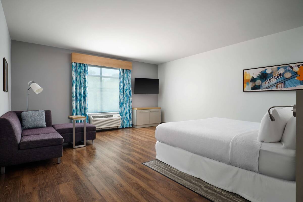A modern hotel room with a sofa, a small table, a bed, a TV, and a window with curtains.