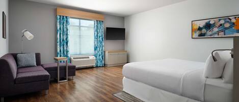 Una habitación de hotel moderna con sofá, una mesita, cama, televisión y ventana con cortinas.