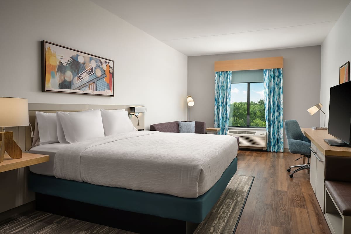 A hotel room with a large bed, a desk with a computer, a TV, and a window with curtains.