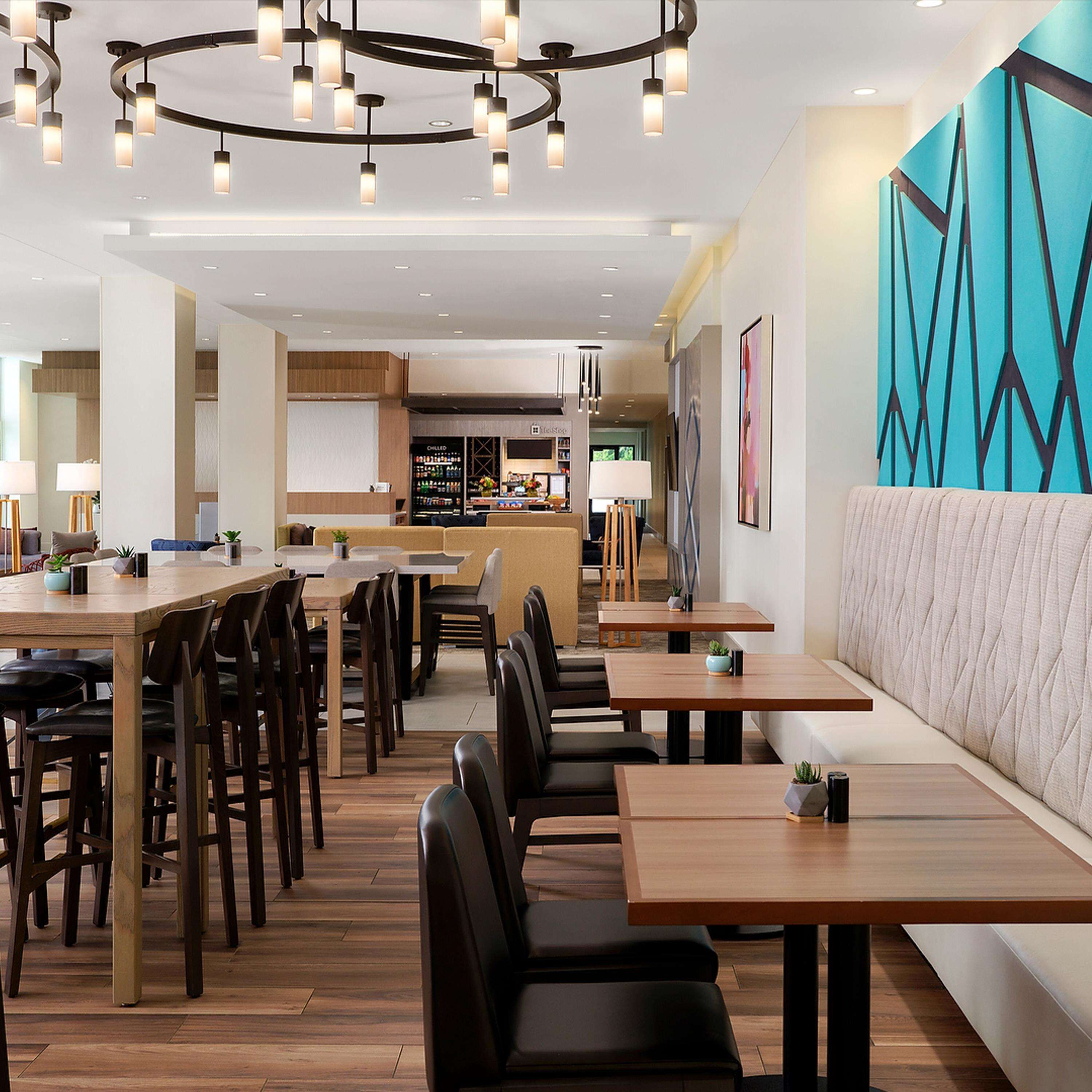 A modern restaurant with wooden tables and chairs, a bar area, and a colorful wall mural.