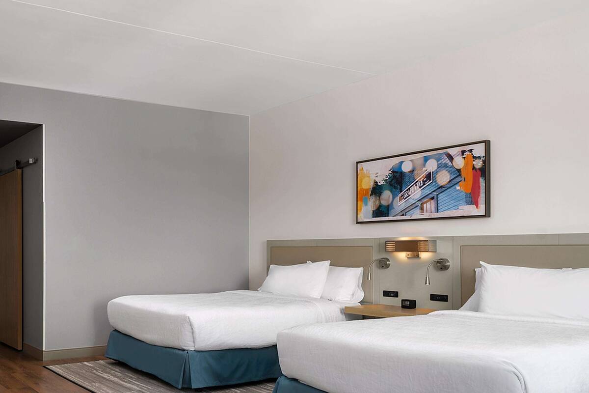 A hotel room with two beds, a wooden floor, a gray rug, and a painting on the wall.