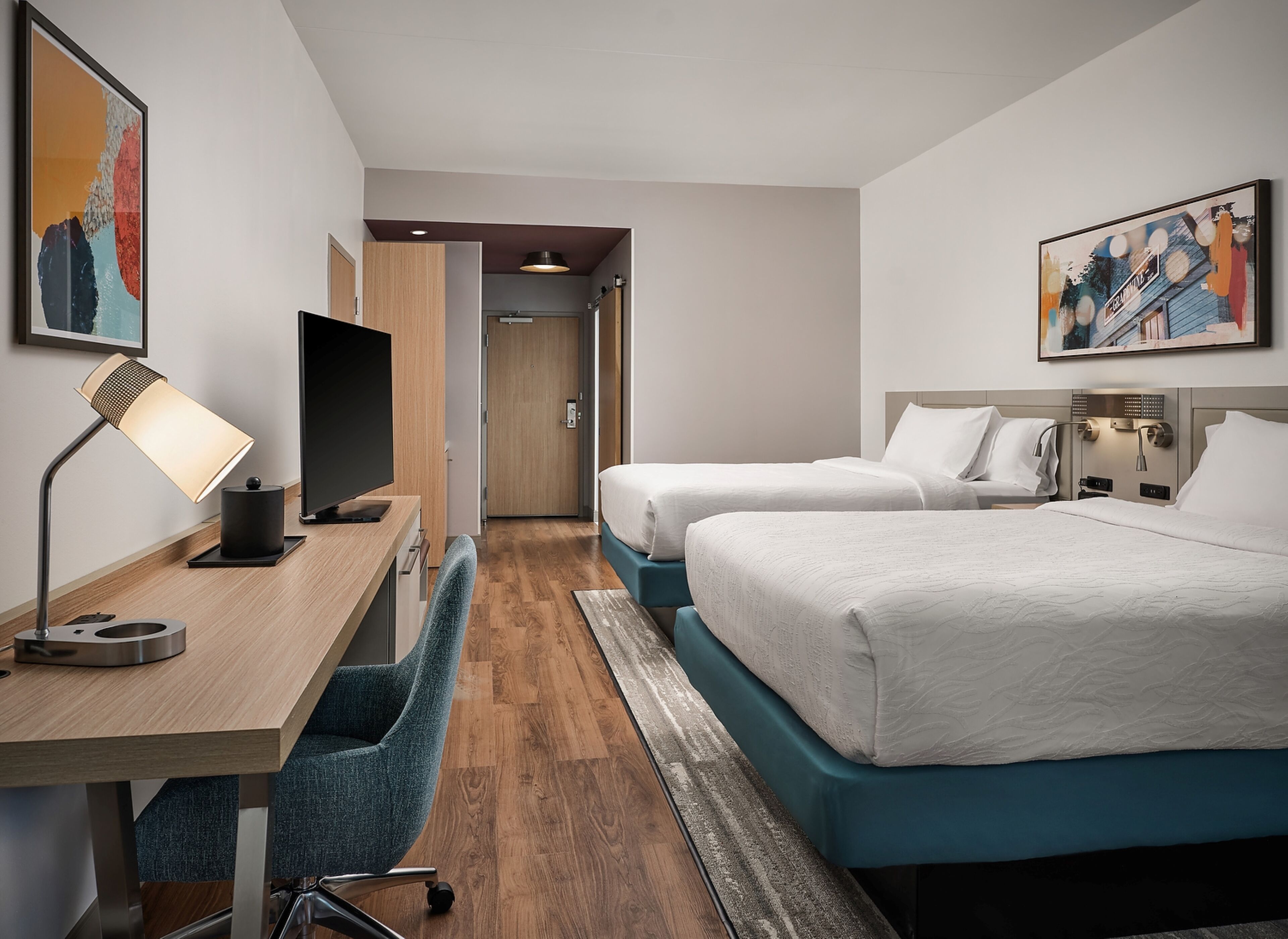 Een hotelkamer met een bureau, twee bedden, een televisie en kunstwerken aan de muren.