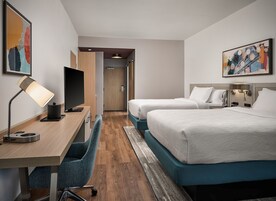 Una habitación de hotel con escritorio, dos camas, televisión y obras de arte en las paredes.