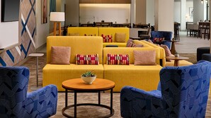 A modern hotel lobby with a yellow sofa set, blue patterned chairs, a round coffee table, and a central chandelier. - Hilton Garden Inn Grapevine at Silverlake Crossing (Grapevine)