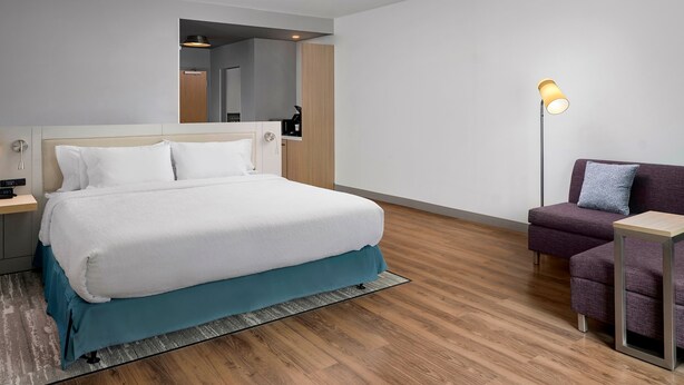 A modern hotel room with a large bed, a sofa, a floor lamp, and a small table. - Hilton Garden Inn Grapevine at Silverlake Crossing (Grapevine)