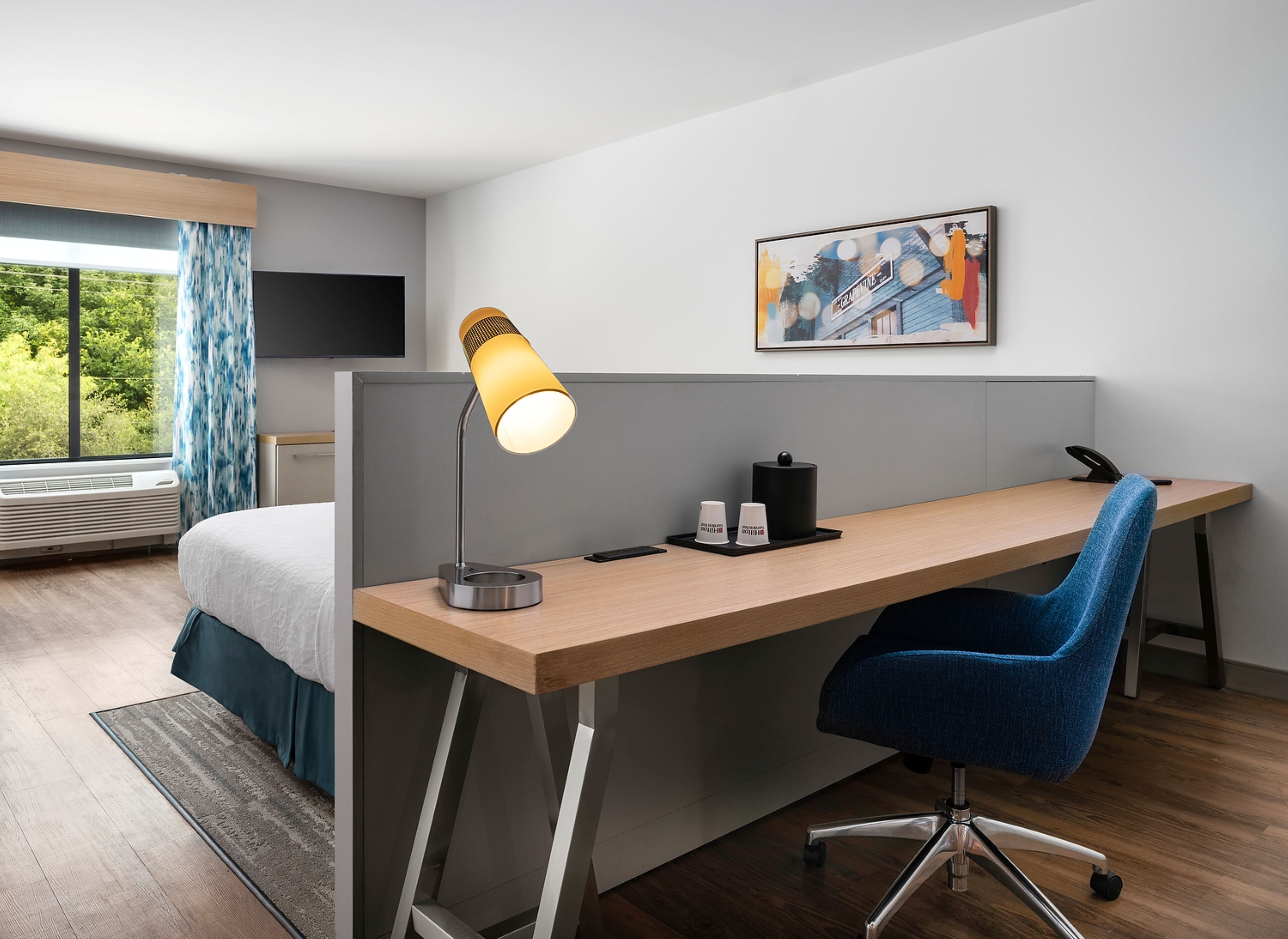 Ein modernes Hotelzimmer mit Schreibtisch aus Holz, blauem Bürostuhl, Schreibtischlampe, Kaffeemaschine und Flachbildfernseher.