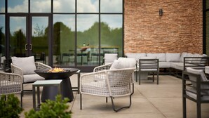 An outdoor patio area with wicker furniture, a fire pit, and a view of the outdoors through large windows. - Hilton Garden Inn Grapevine at Silverlake Crossing (Grapevine)