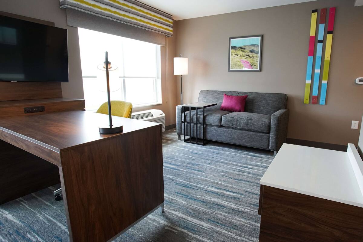 Ein modernes Hotelzimmer mit Flachbildfernseher, Schreibtisch mit Lampe, grauem Sofa mit rosa Kissen und einem farbenfrohen abstrakten Kunstwerk an der Wand.