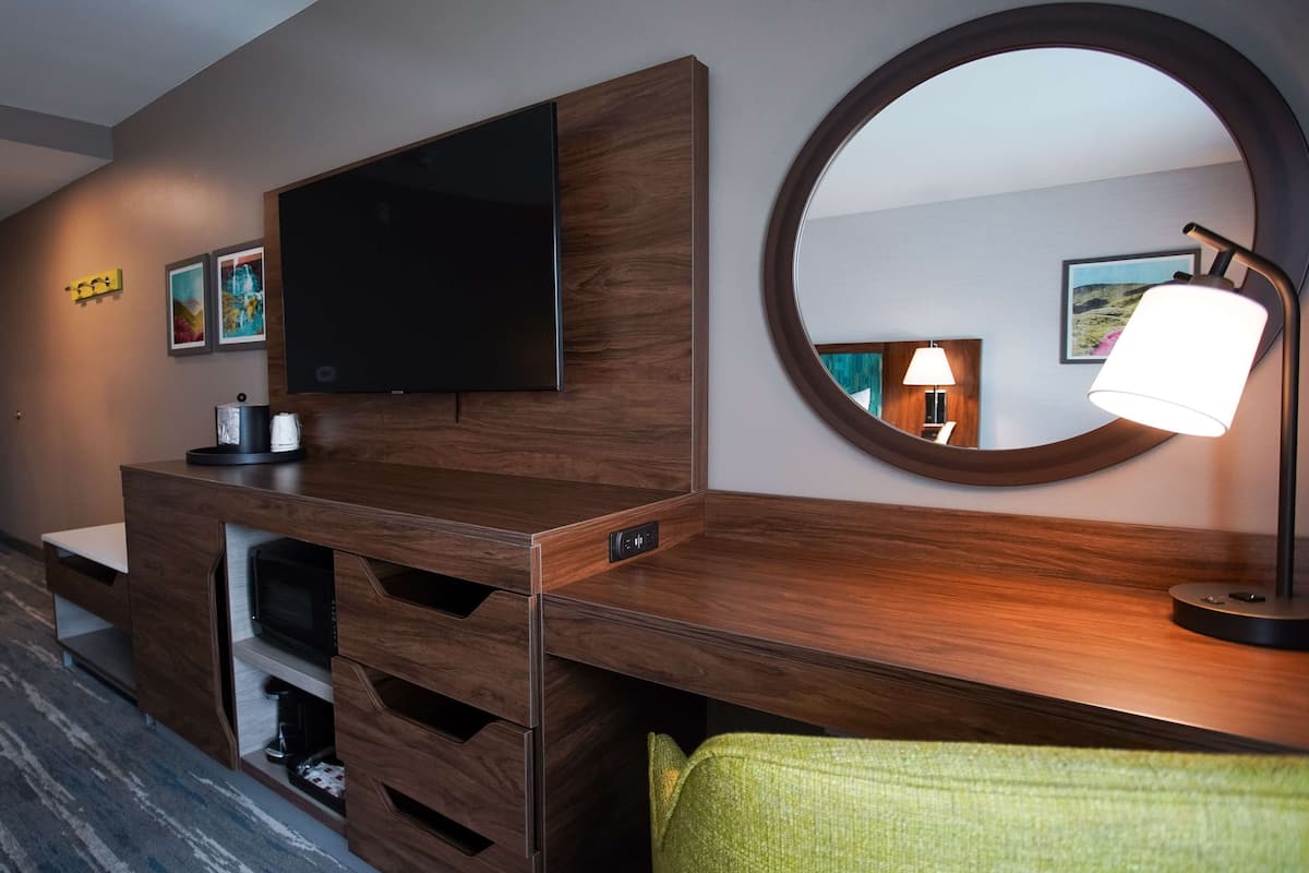 Ein modernes Hotelzimmer mit Schreibtisch aus Holz, einem Flachbildfernseher, einem Spiegel und einer Lampe.