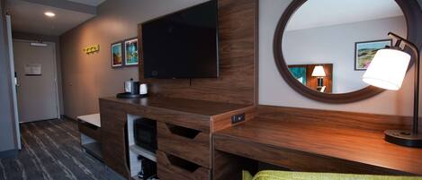 A modern hotel room with a wooden desk, a flat-screen TV, a mirror, and a lamp.