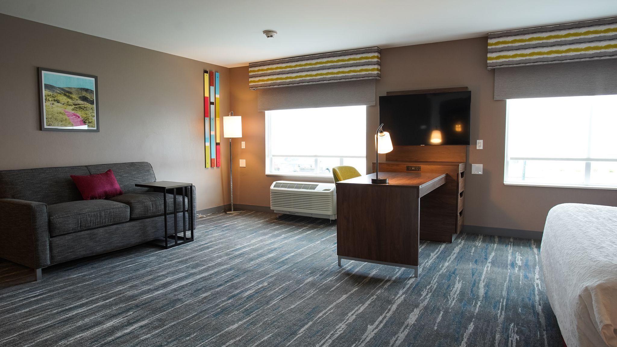 A hotel room with a gray sofa, a desk with a chair, a flat-screen TV, and a large window.